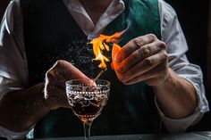 a man in an apron is holding a glass with something on it and fire coming out of the top