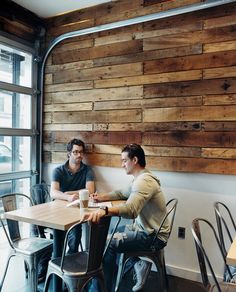 two men sitting at a table in front of a window with wood paneling on the wall