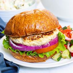 a chicken sandwich with lettuce, tomato and onion on a white plate next to a bowl of salad