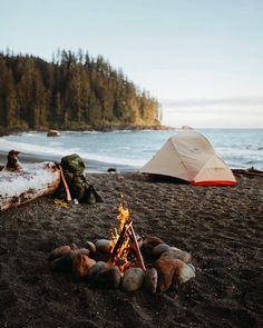 there is a campfire on the beach next to some tents and trees in the background