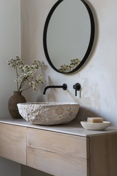 a bathroom sink sitting under a round mirror next to a vase with flowers in it