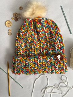 a multicolored knitted beanie sits next to knitting needles and yarn