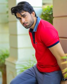 a man in a red shirt and blue pants leaning against a wall with his hands on his hips