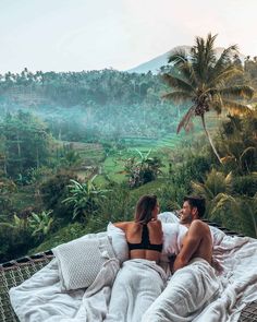 two people sitting on a bed in the middle of a jungle with lush green fields and palm trees