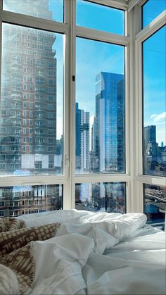 a bed with white sheets and pillows in front of two large windows overlooking the city