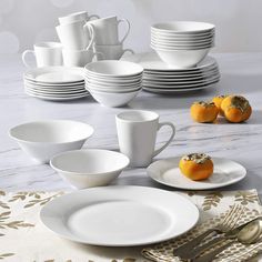 a table set with white dishes, silverware and oranges on the tablecloth