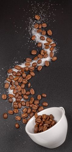 coffee beans spilled out of a white bowl on a black surface next to some sugar