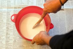 a person using a spatula to mix milk in a red bowl on top of newspaper