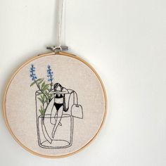 a embroidery hanging on a wall with a plant in a vase and some blue flowers