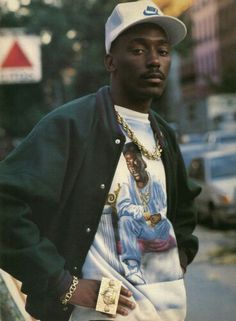 a man in a baseball cap and jacket standing next to a street with his hands in his pockets