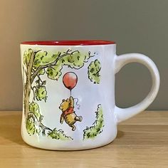 a winnie the pooh coffee mug on a wooden table with trees and balloons painted on it