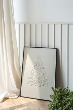 a framed art piece sitting on top of a wooden floor next to a window with white curtains