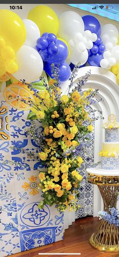 a table topped with a cake covered in yellow and blue balloons next to a vase filled with flowers