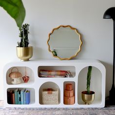 a white shelf with books and plants on it next to a mirror, potted plant and other items