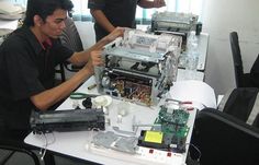 two men working on an electronic device at a table with other people in the background