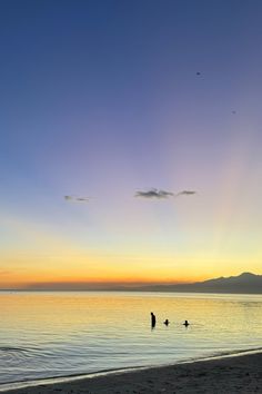 the sun is setting at the beach with people swimming in the water