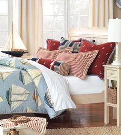 a bed with red and blue pillows on it next to a white headboard in front of a window