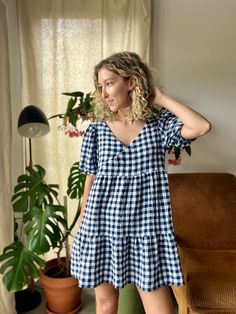 a woman in a blue and white checkered dress is standing by a window with her hands on her head