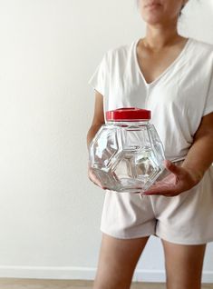 a woman holding a glass jar with red lid in front of her face and wearing white shorts