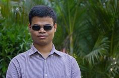 a young man wearing sunglasses standing in front of some trees and bushes with his hand on his hip