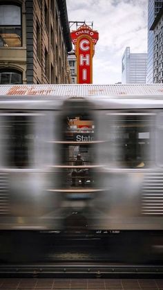 a silver train traveling past tall buildings on a city street in front of a neon sign