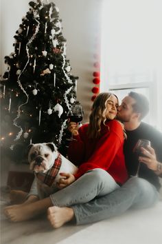 a man and woman sitting on the floor next to a christmas tree with a dog