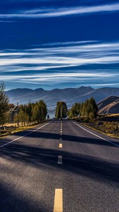 an empty road with trees and mountains in the backgrouds, under a blue sky