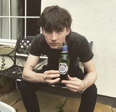 a young man sitting on a bench holding a beer bottle in his right hand and looking at the camera