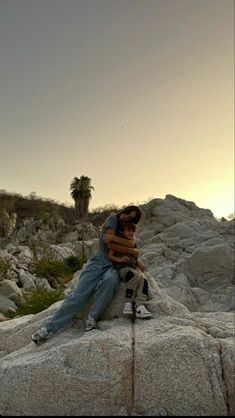 a man sitting on top of a large rock next to a palm tree in the distance