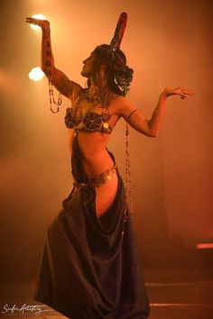 a belly dancer is performing on stage with her hands in the air and lights behind her