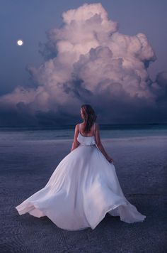 a woman in a white dress is standing on the beach at night under a cloudy sky