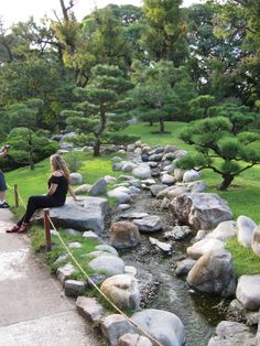 two people sitting on rocks near a small stream in a park with green grass and trees