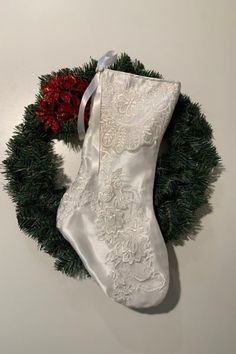 a white christmas stocking hanging on a wall next to a green wreath with two red poinsettis