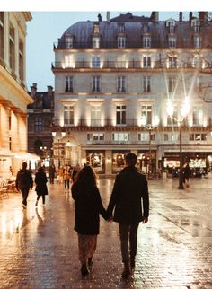 two people holding hands walking down the street in front of an old building at night