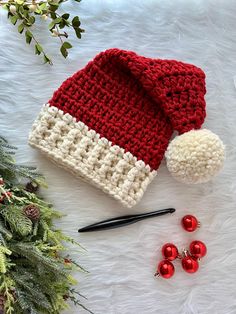 a red and white knitted hat next to christmas decorations on a furry surface with greenery