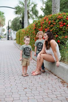 a woman and two children are sitting on the curb by some bushes with red flowers