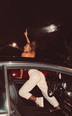 two people sitting in the back of a car at night, one person waving to the sky