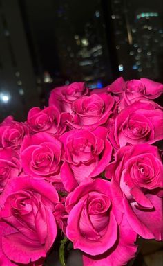 a bunch of pink roses in a vase on a table with city lights behind them