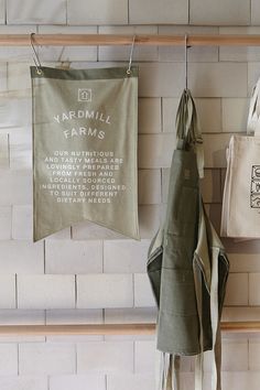 two aprons hanging from hooks on a brick wall next to other kitchen towels and bags