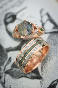 A matching wedding ring set from Staghead Designs sits on an antique bird illustration. First is our "Lucy In The Sky" design engagement ring in 14K rose gold with a hexagon cut moss agate center stone, diamond halo & accents, & moss inlays. Below is "Rio" in 14K rose gold with a centered crushed moldavite inlay & a damascus steel inlay on either side. These rings are fully customizable & can be made with a wide variety of unique materials & stones. Make Wedding Rings, Moss Agate Matching Rings, Wedding Rings With Green Stones, Libra Engagement Ring, Moss Agate Band Ring, Matching Moss Agate Wedding Rings, Moldavite Engagement Ring, Miss Agate Wedding Ring, Womens Engagement Rings Unique