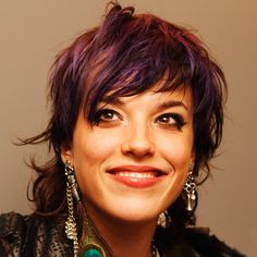 a close up of a person with purple hair and necklaces smiling at the camera