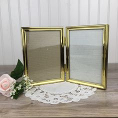 two gold frames sitting on top of a wooden table next to a pink rose and lace doily