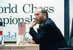 a man sitting at a desk with an american flag in front of him and the world chess championship banner behind him