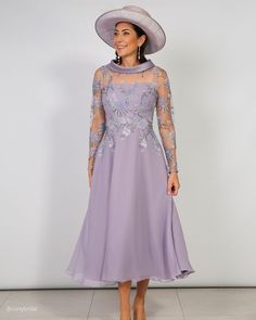 a woman wearing a purple dress and hat with long sleeves, standing in front of a white wall