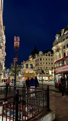 people are walking down the street in front of some buildings at night with lights on