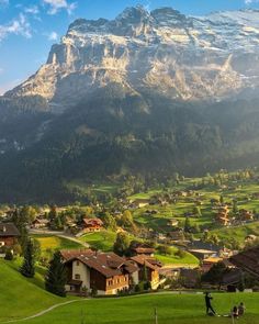 people are standing on the grass in front of some mountains and houses with snow capped peaks