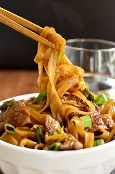 a white bowl filled with noodles and meat on top of a wooden table next to chopsticks