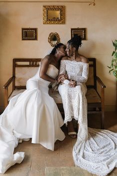 two brides sitting on a bench kissing each other in front of a wall with pictures