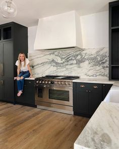 a woman sitting on the counter in a kitchen with black cabinets and marble backsplash