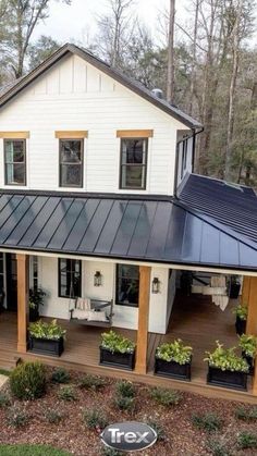 a white house with black metal roof and plants on the front porch, surrounded by trees